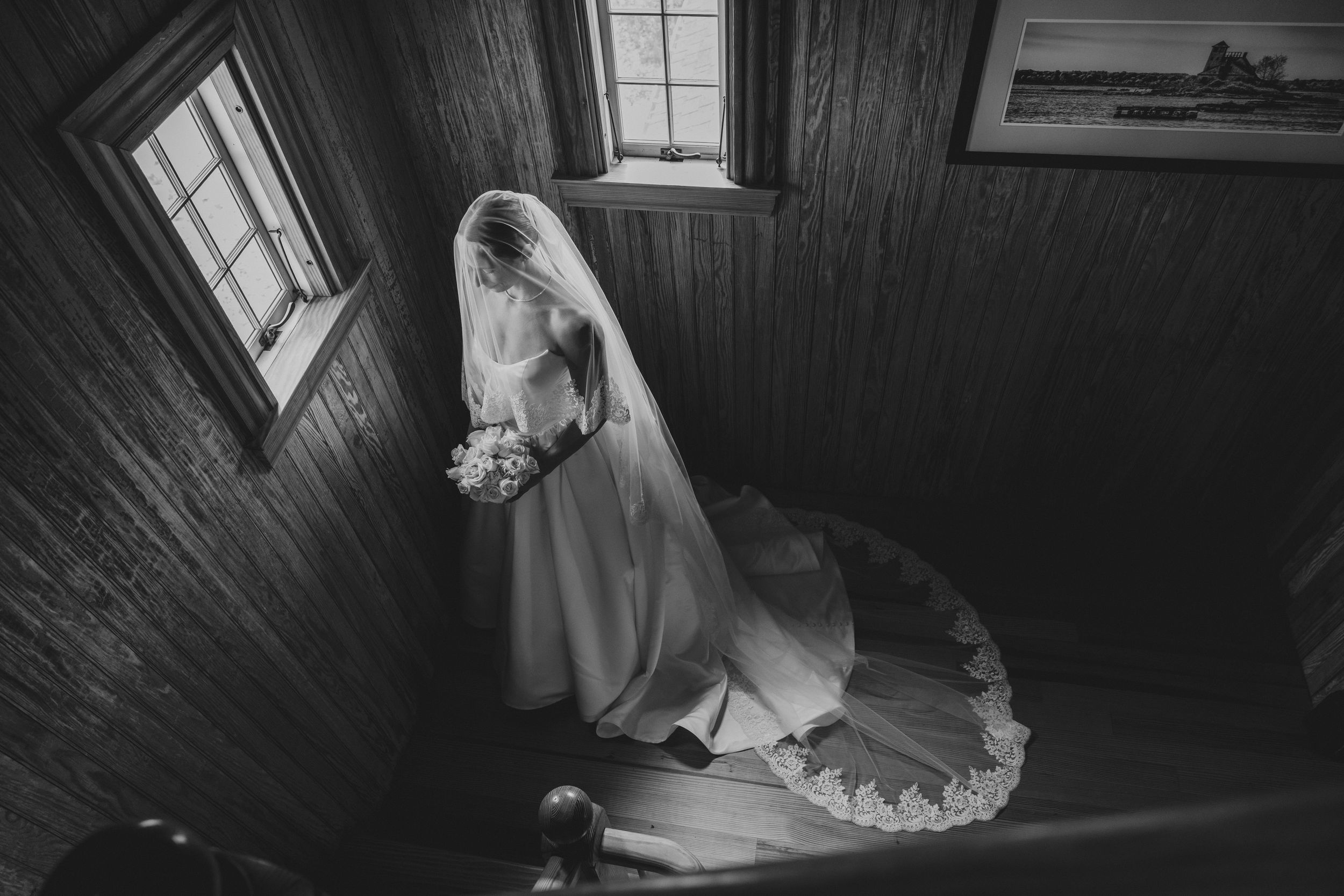 Black and white photo of a bride in a white gown and veil holding a bouquet on a wooden staircase.