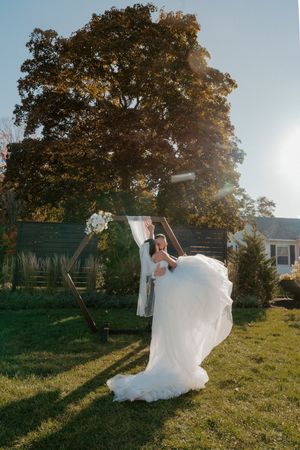 Groom lifting bride in flowing white gown outdoors, sunlit backyard wedding with large tree backdrop.