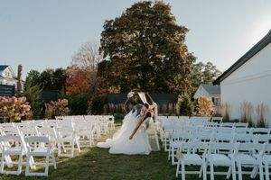 Couple sharing a romantic dip kiss at an outdoor wedding ceremony with white chairs and autumn trees.