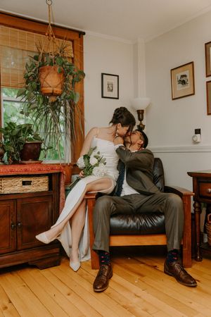 Bride and groom sharing a kiss while seated in a cozy, plant-filled room.