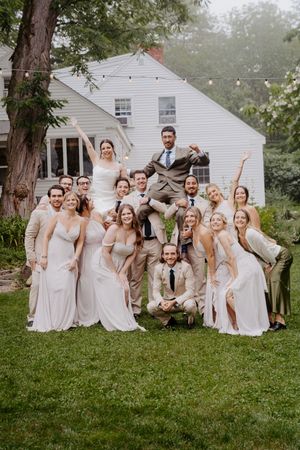 Joyful wedding party posing together outside a white house surrounded by greenery.