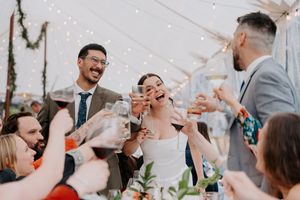 Bride and groom laughing during wedding toast with guests raising glasses under a tent with string lights.