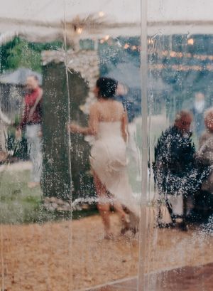 Bride in white dress dancing at outdoor wedding reception viewed through rain-speckled clear tent panels.