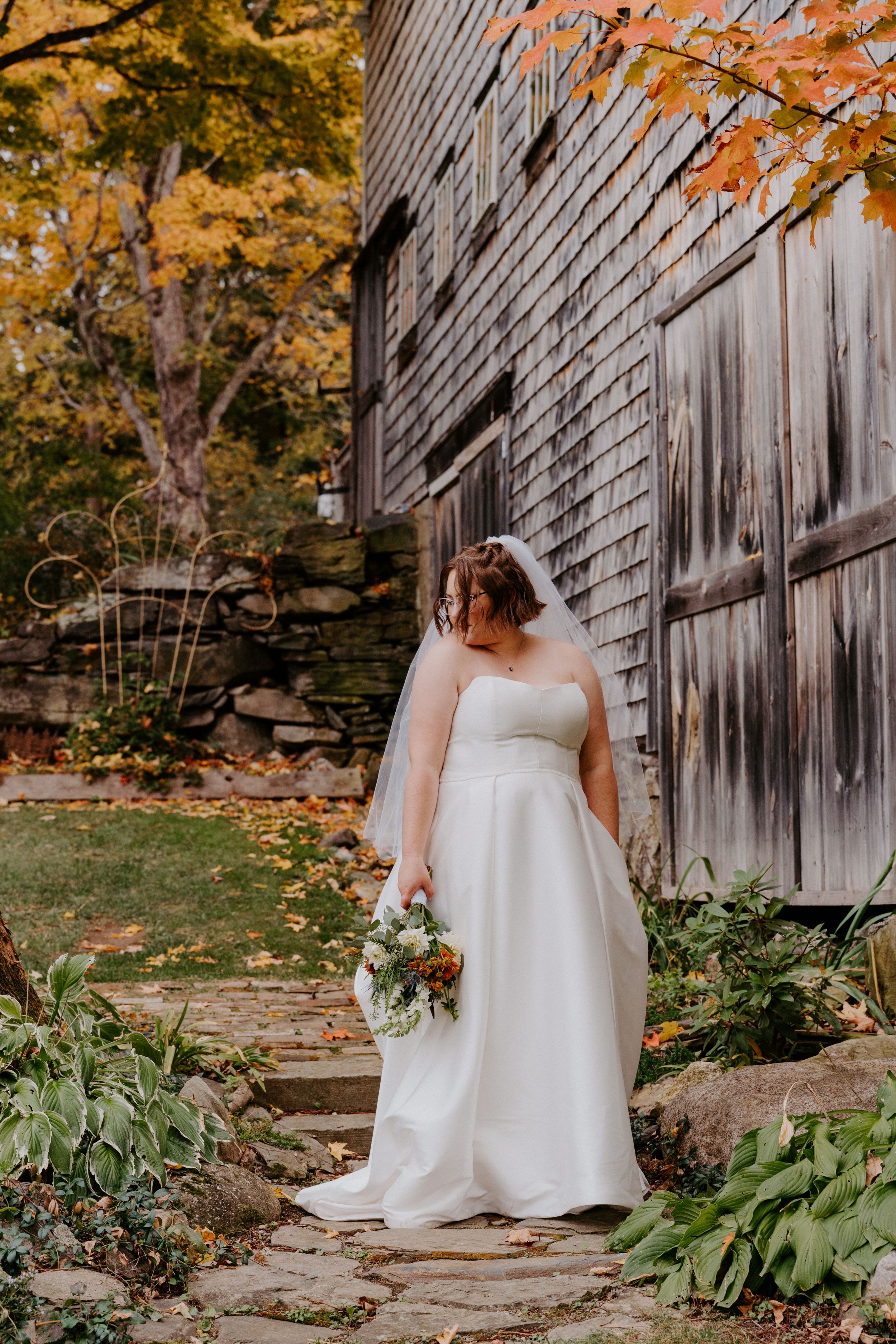 A bride in a classic white dress holds a bouquet while standing beside a weathered wooden barn in fall.
