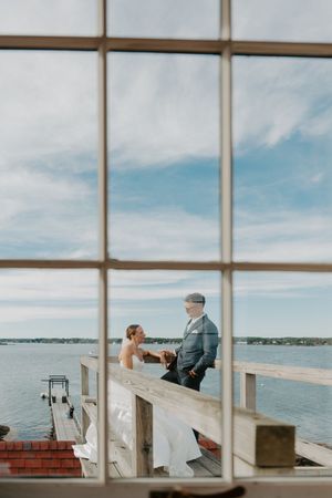 Bride and groom share a romantic moment on a waterfront deck, viewed through large windows with blue sky backdrop.