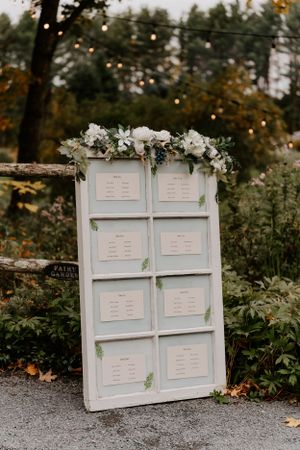 White vintage door decorated with flowers displays wedding seating chart in outdoor garden setting.