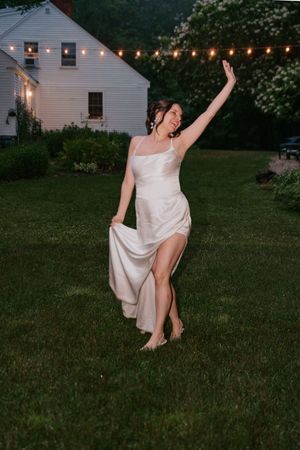 Joyful bride in white satin gown dances barefoot on lawn at twilight wedding reception.
