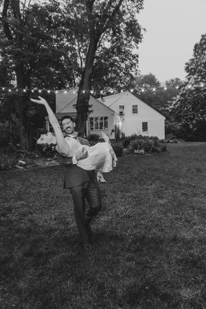 Black and white photo of groom lifting bride joyfully on lawn at outdoor wedding.