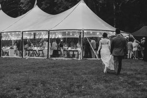 Black and white photo of a bride and groom walking toward a festive wedding tent with string lights.