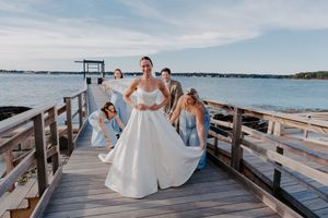 Bride in white ball gown poses on wooden dock with bridesmaids in blue by waterfront on sunny day.