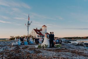 Outdoor wedding ceremony on rocky Maine coastline with white lighthouse building and American flag at golden hour sunset.