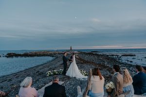 Couple exchanges vows at a scenic coastal wedding ceremony at dusk with guests watching near a rocky shoreline.