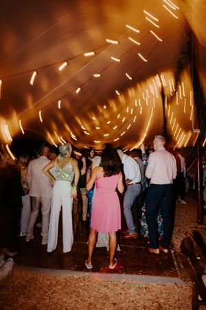 Guests dancing under warm glowing lights inside a festive wedding tent at night.