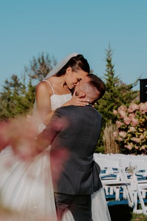 Bride and groom share a romantic kiss outdoors at a wedding ceremony with white chairs and pink flowers.