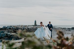 A bride and groom walk hand-in-hand along a rocky New England coastline with a lighthouse in the background.