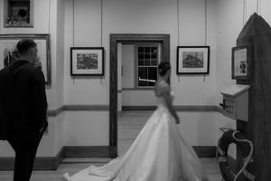 Black and white wedding photo of bride in flowing gown and groom in gallery hall with framed artwork on walls.