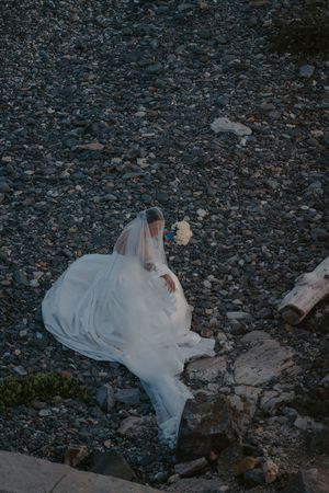 Bride in white gown and veil sitting on rocky shoreline holding bouquet, aerial view at dusk.