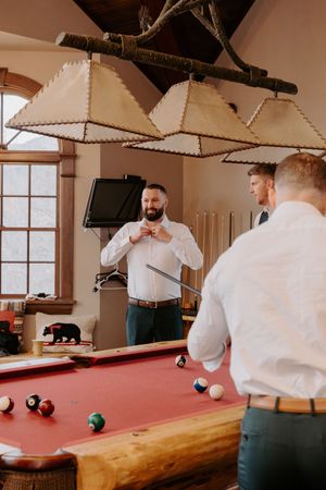 Two groomsmen in white shirts playing pool on a red billiard table beneath rustic log-framed pendant lights indoors.