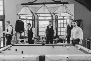 Black and white photo of groomsmen gathered around a pool table in a rustic wedding venue billiard room.