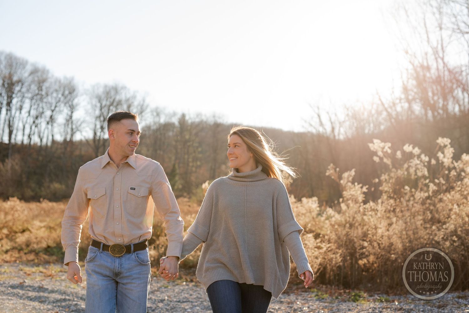 A couple takes a romantic stroll through a sunlit field with golden grasses and bare trees in the background.