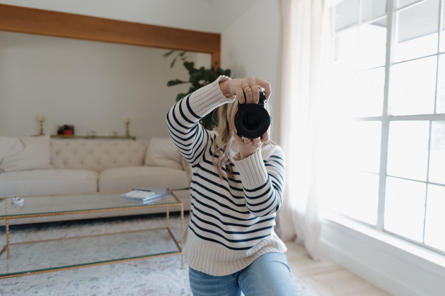 A person wearing a striped sweater takes a photograph near a bright window in a minimalist living room.