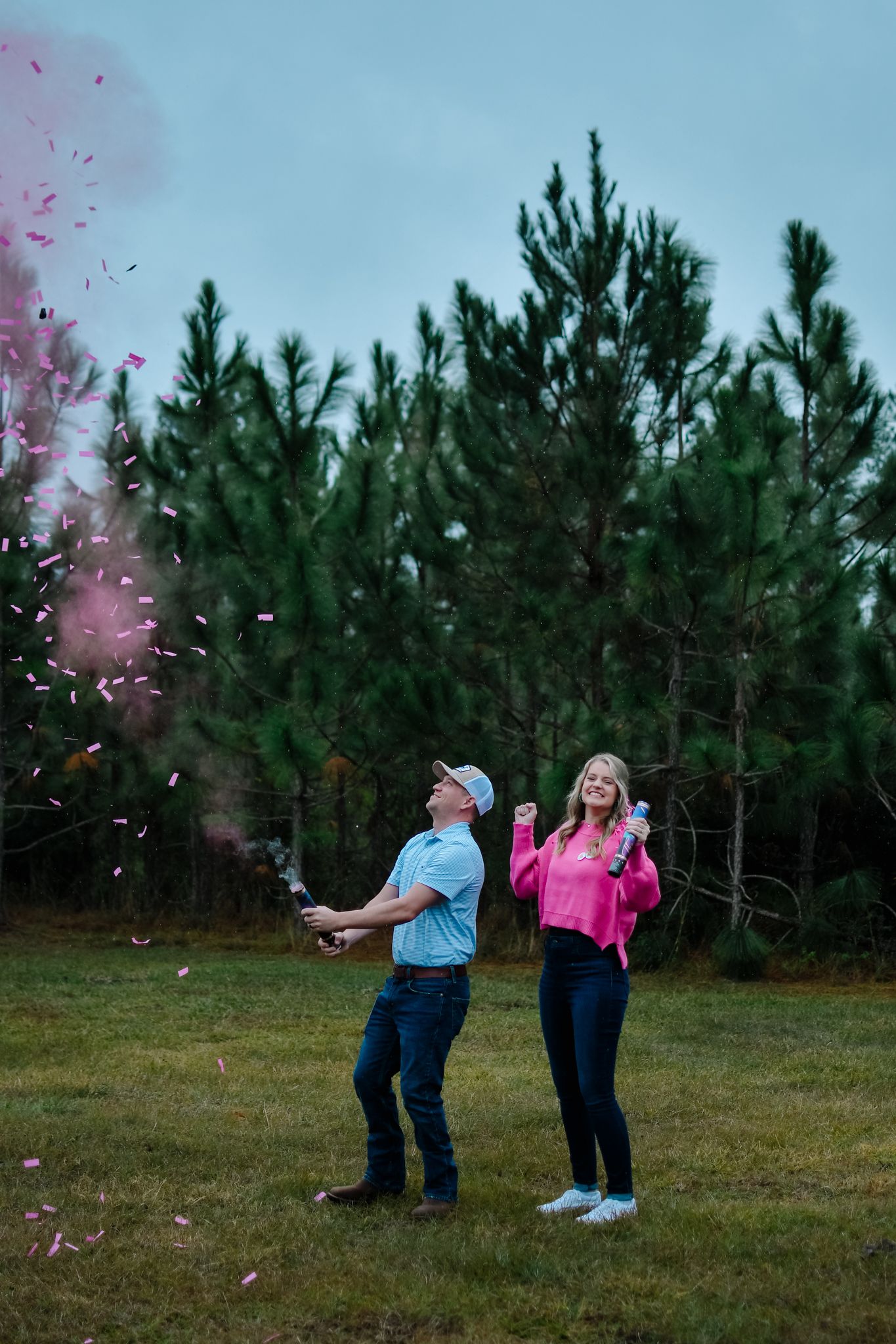 Two people throw pink confetti while standing in a grassy field with evergreen trees.