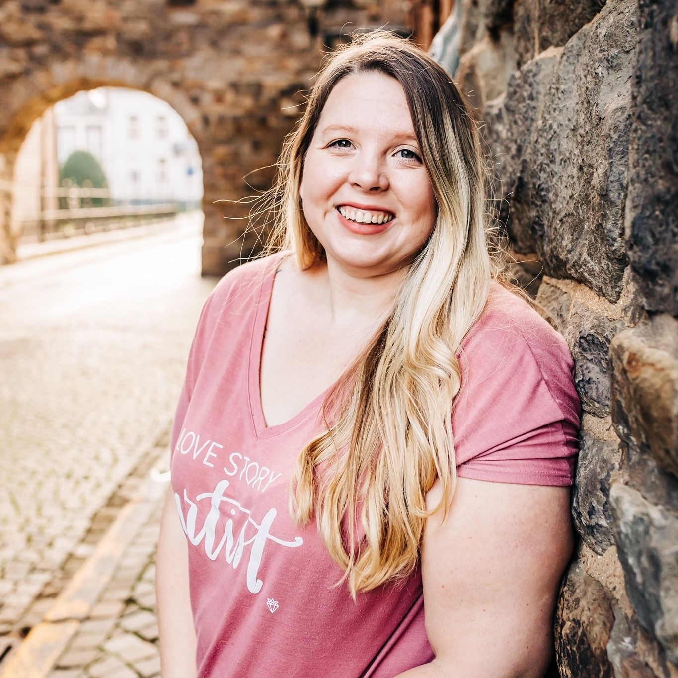 Woman in pink t-shirt smiling near stone archway on sunny day.