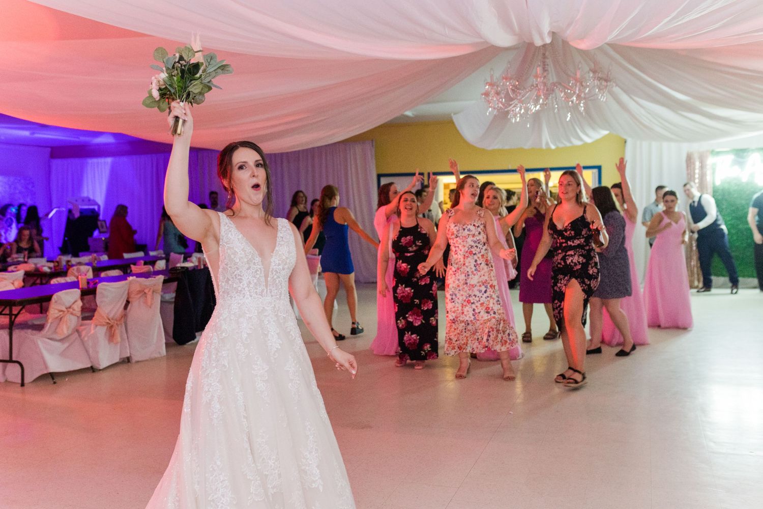 A joyful wedding celebration with guests dancing under pink and purple mood lighting.