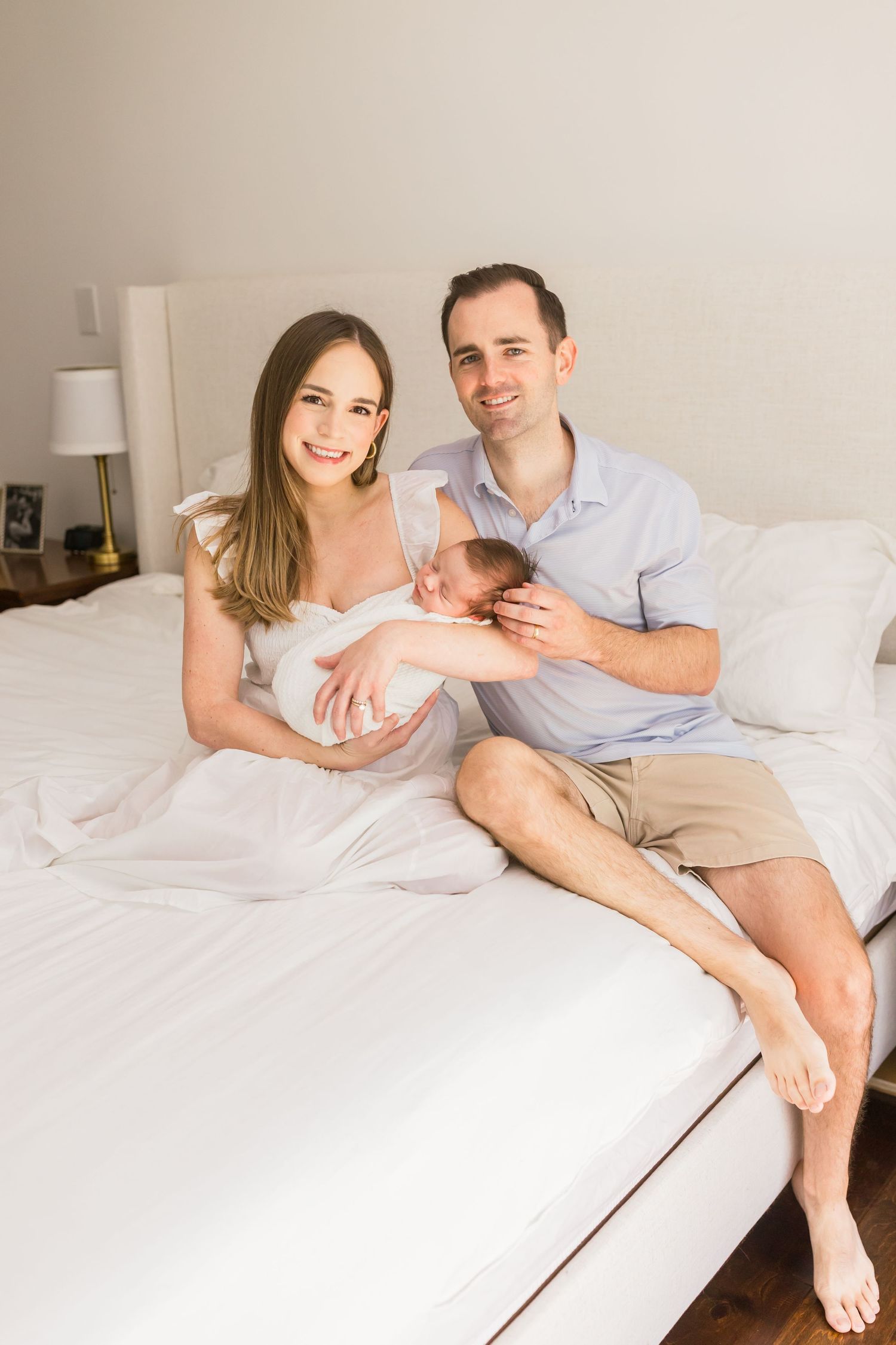 A couple poses with their newborn baby while sitting on a white bed with bedside lamp visible.