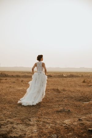 A bride in a flowing white dress stands alone in a desert landscape during golden hour.