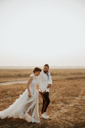 A wedding couple walks hand in hand across an arid landscape at sunset.