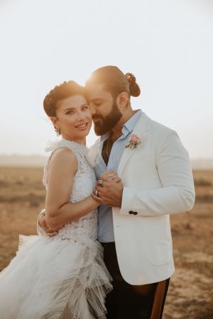 A joyful wedding couple embraces in formal attire against a desert landscape at sunset.