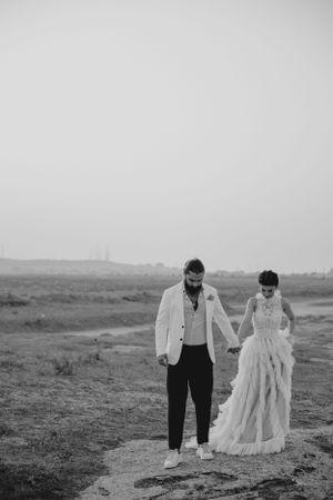 A couple in wedding attire walking hand in hand across a barren landscape in black and white.