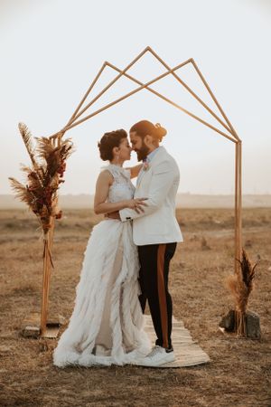 Newlyweds embrace under a geometric arch with dried floral arrangements at sunset.