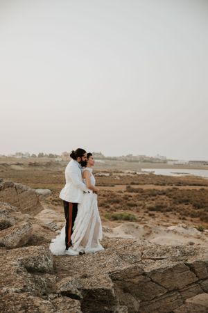 A couple in wedding attire stands together on rocky coastal cliffs at sunset.