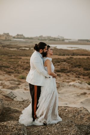 A couple in wedding attire embraces while overlooking a coastal landscape at sunset.