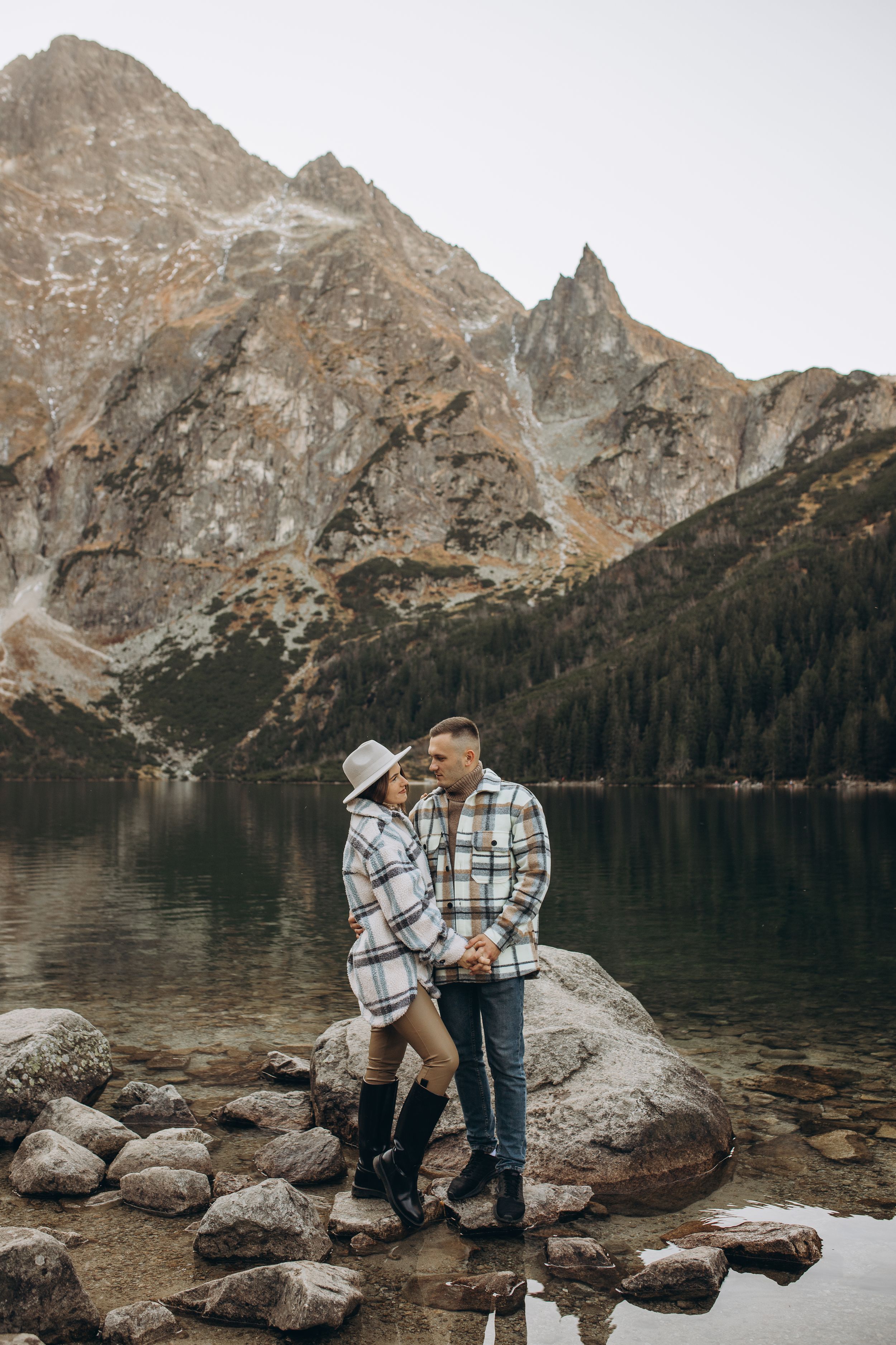 Two people in coordinated outfits stand on rocks beside a serene mountain lake.