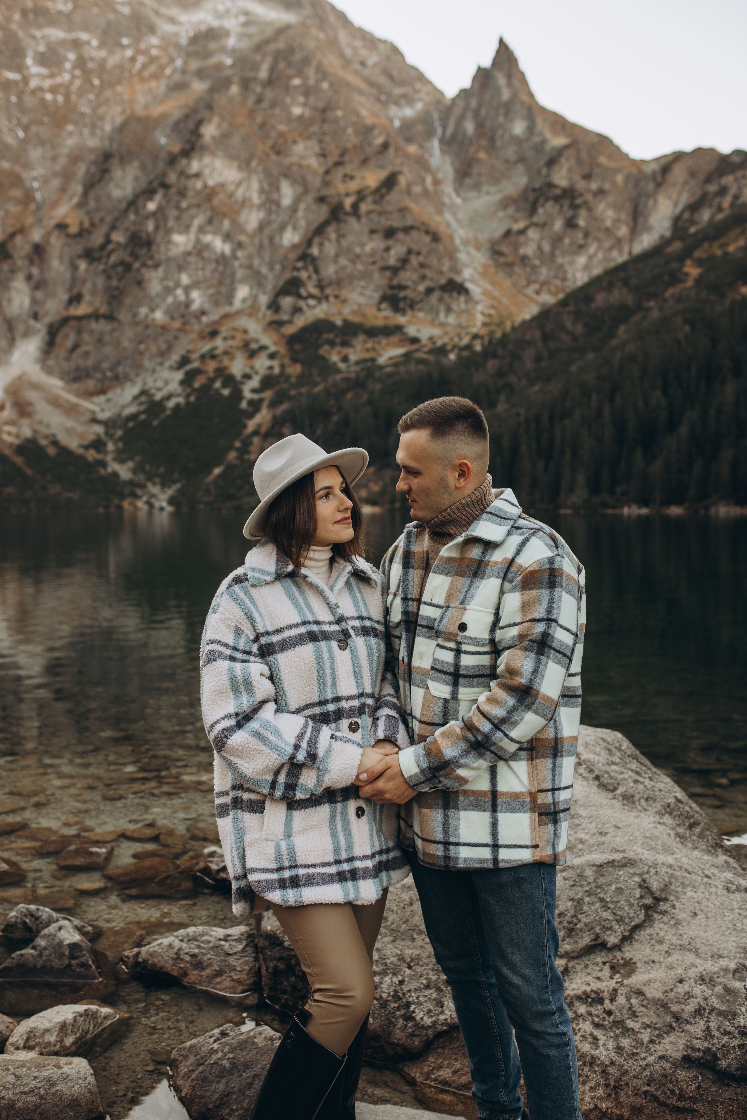 A couple wearing matching plaid coats share an intimate moment by a mountain lake.