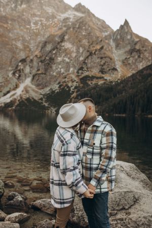 A couple in matching plaid jackets holds hands by a scenic mountain lake.