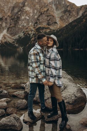 A loving couple in coordinated plaid outfits embrace on rocks by an alpine lake.