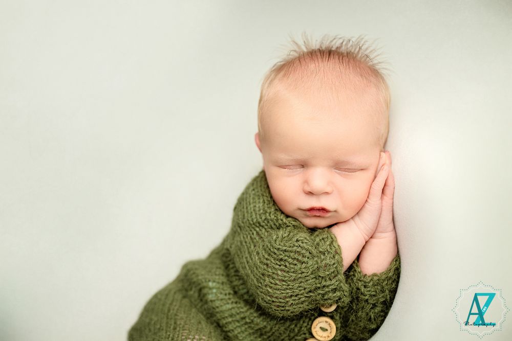 Peaceful newborn wrapped in green knit wear resting face on hands.