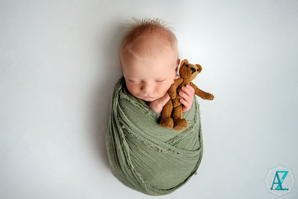 Sleeping newborn wrapped in sage green fabric with small brown teddy bear.