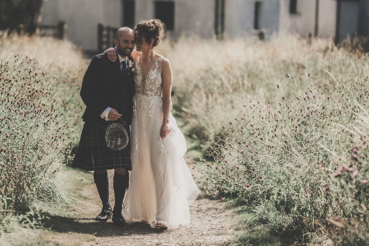 A couple in wedding attire embraces on a path surrounded by tall grass and wildflowers.