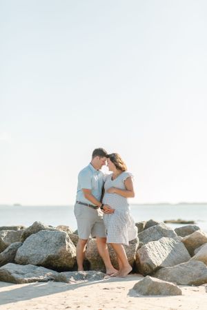 Expecting couple stands together on rocky beach during coastal maternity photoshoot in Hilton Head, South Carolina by local photographer.
