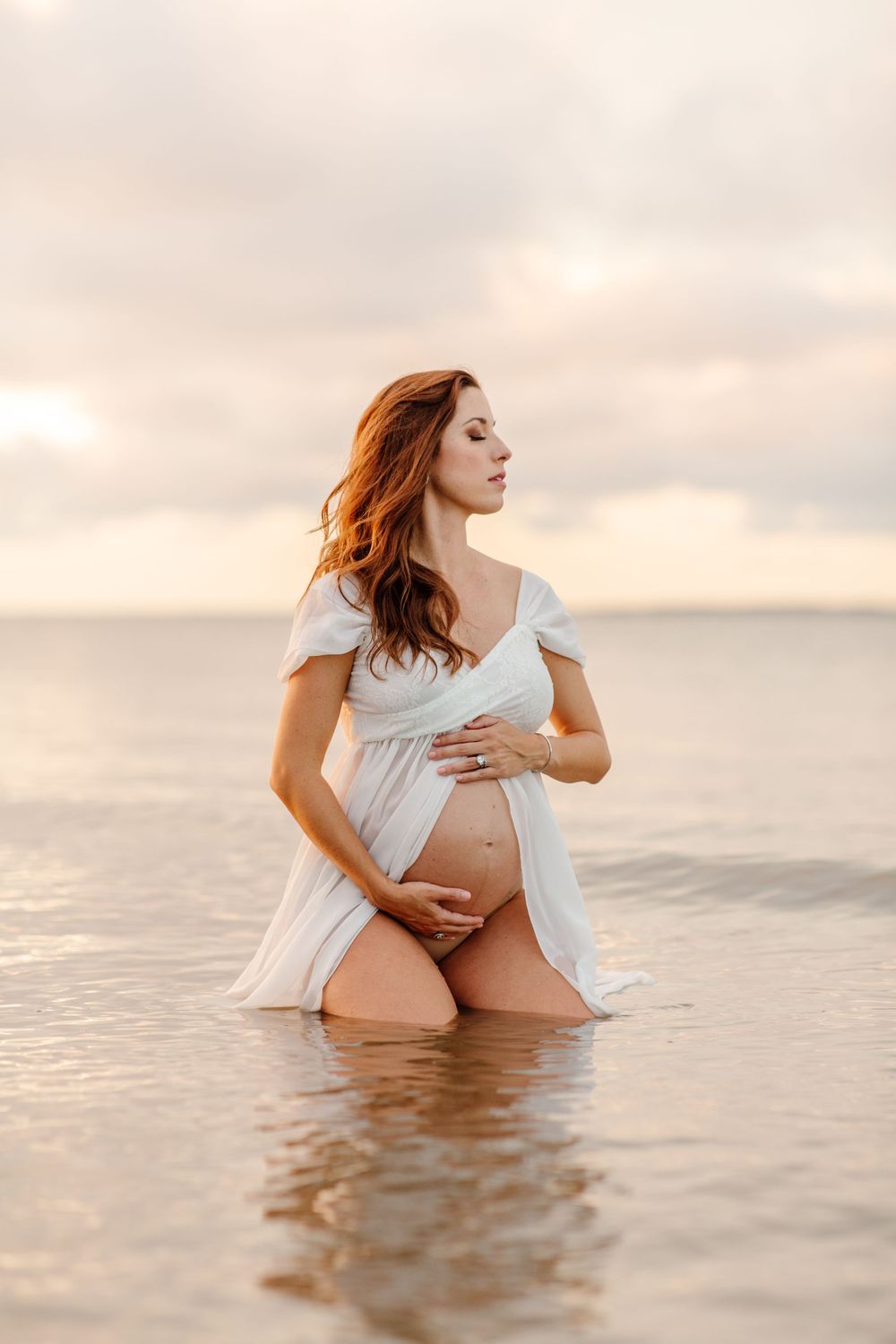 An artistic maternity photo of a woman in white kneeling in ocean water during golden hour in Mitchelville beach in Hilton Head, SC.