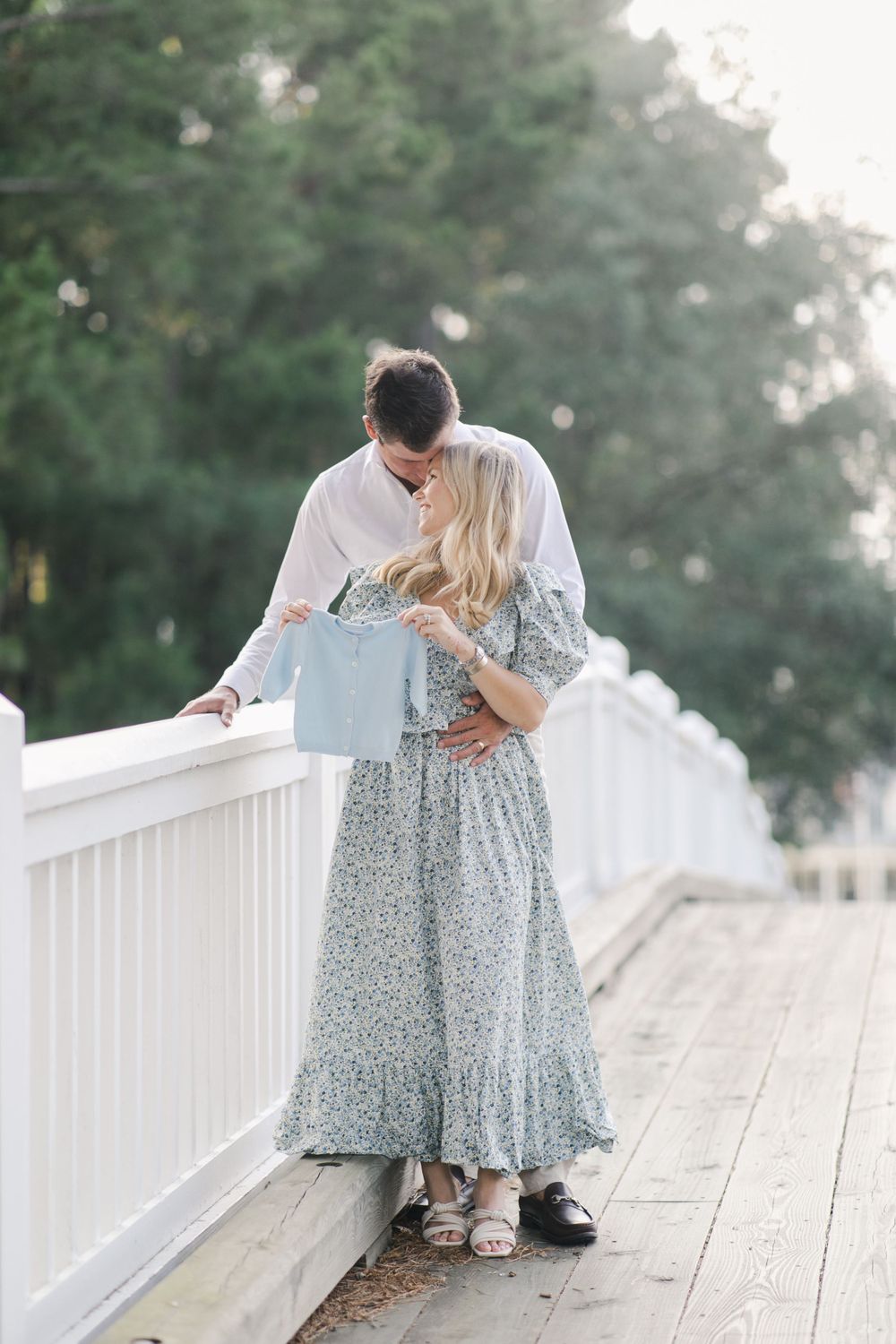 Couple announcing baby embraces on a white wooden deck with lush greenery in the background in Old Field, Bluffton, SC.