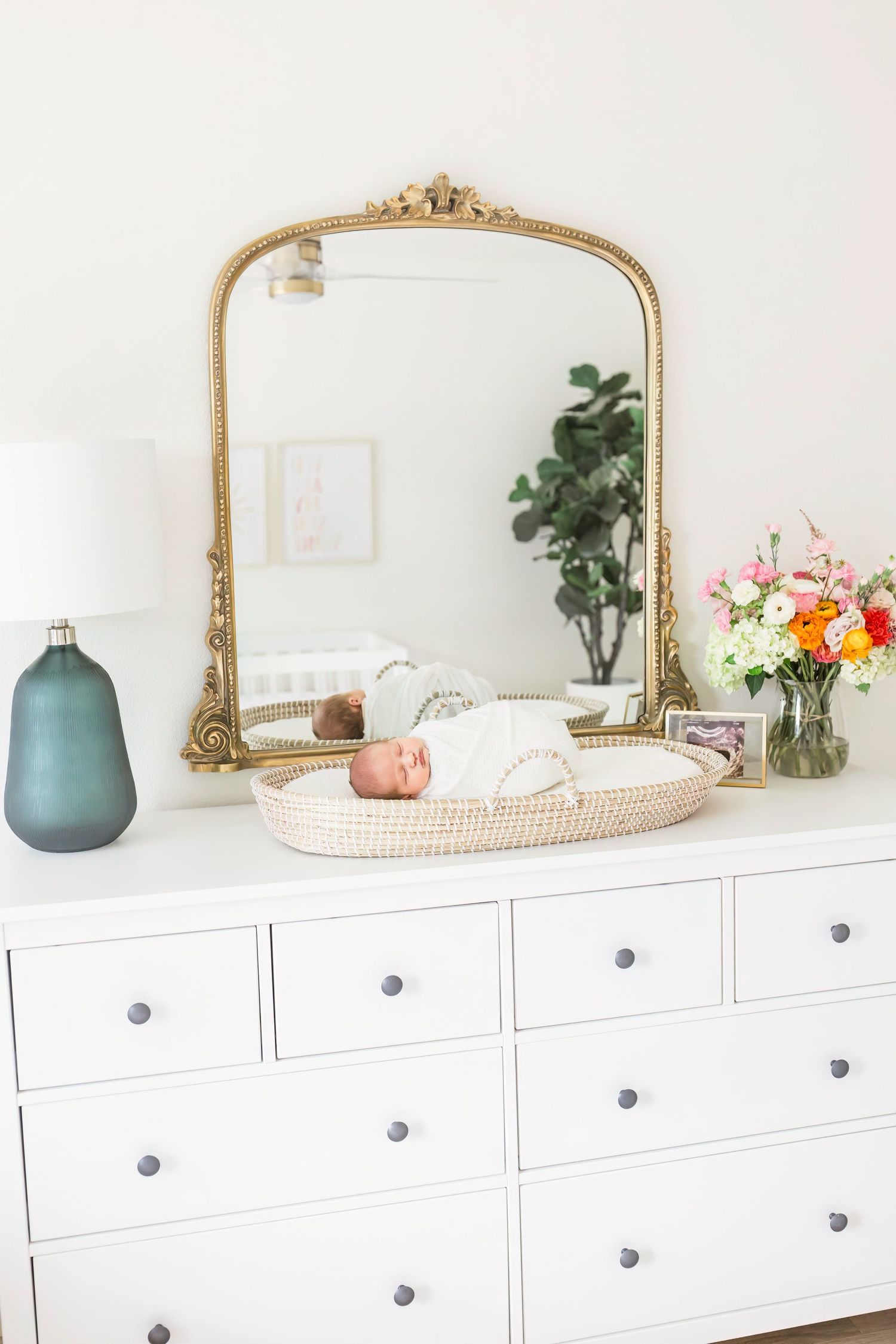 Ornate gold mirror on white dresser with teal vase and floral arrangement.