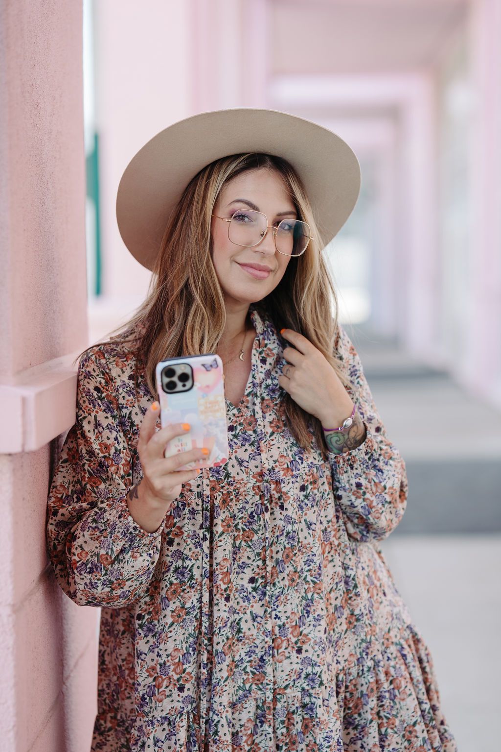 Woman in floral dress and hat posing with phone against pink columns.