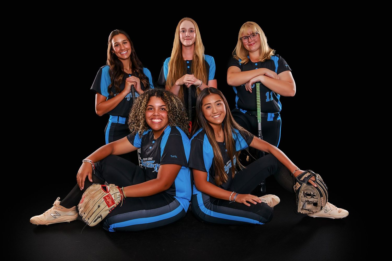 Female softball team members in blue uniforms pose for a team photo on black background.