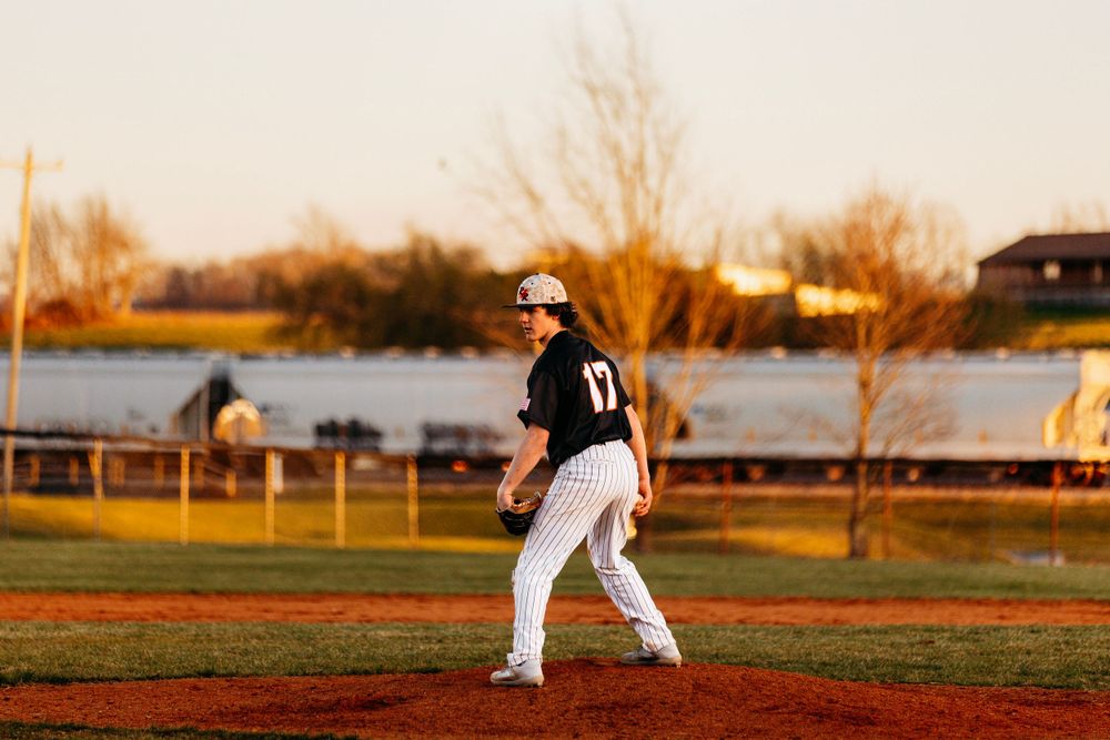 Baseball pitcher number 17 in black uniform walks on mound during a sunset evening game.
